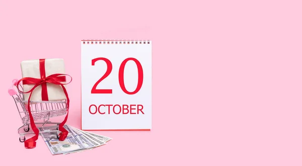 Pudełko prezentów w wózku sklepowym, dolarach i kalendarzu z datą 20 października na różowym tle. — Zdjęcie stockowe