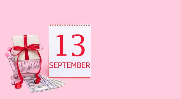 Pudełko prezentów w wózku sklepowym, dolarach i kalendarzu z datą 13 września na różowym tle. — Zdjęcie stockowe
