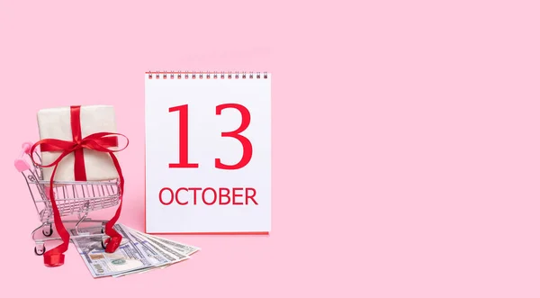 Eine Geschenkbox im Einkaufswagen, Dollars und ein Kalender mit dem Datum des 13. Oktober auf rosa Hintergrund. — Stockfoto