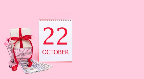 Un coffret cadeau dans un chariot, des dollars et un calendrier avec la date du 22 octobre sur fond rose. — Photo