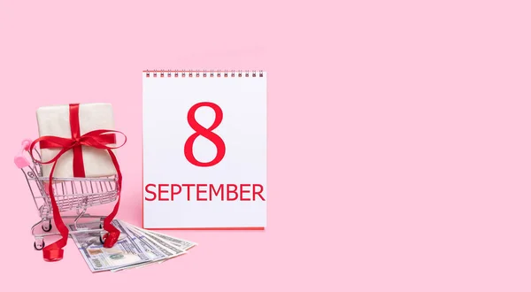 Pudełko prezentów w wózku sklepowym, dolarach i kalendarzu z datą 8 września na różowym tle. — Zdjęcie stockowe