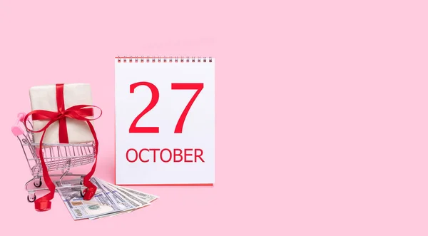 Un coffret cadeau dans un chariot, des dollars et un calendrier avec la date du 27 octobre sur fond rose. — Photo