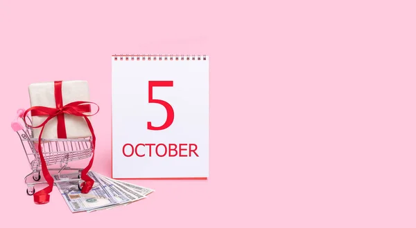 Eine Geschenkbox im Einkaufswagen, Dollars und ein Kalender mit dem Datum des 5. Oktober auf rosa Hintergrund. — Stockfoto