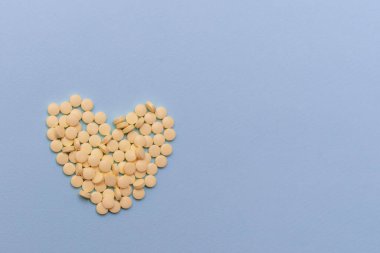 Mavi arka planda kalp şeklinde bir yığın sarı hapın tıbbi geçmişi, fotokopi alanı, üst görünüm, düz yatış. Mavi arka planda sarı haplardan yapılmış kalp, kalp hastalıkları için ilaçlar.