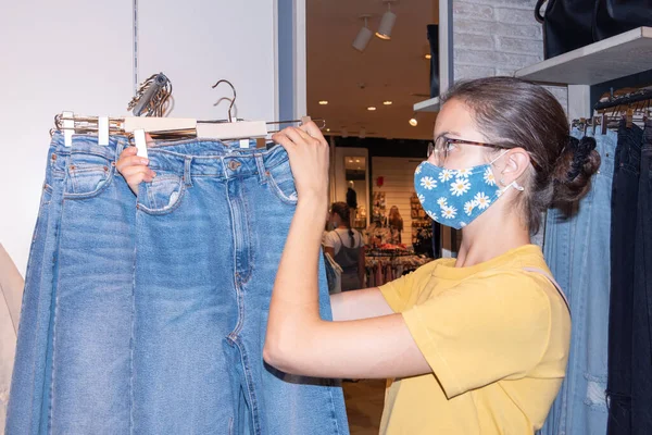 Girl in a protective mask choosing jeans in a boutique. Safe shopping in stores concept. Seasonal sale concept