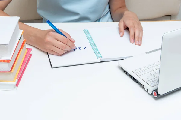 Estudiante sosteniendo un bolígrafo, escribiendo en un cuaderno la tarea, sentada frente a un portátil y libros. — Foto de Stock