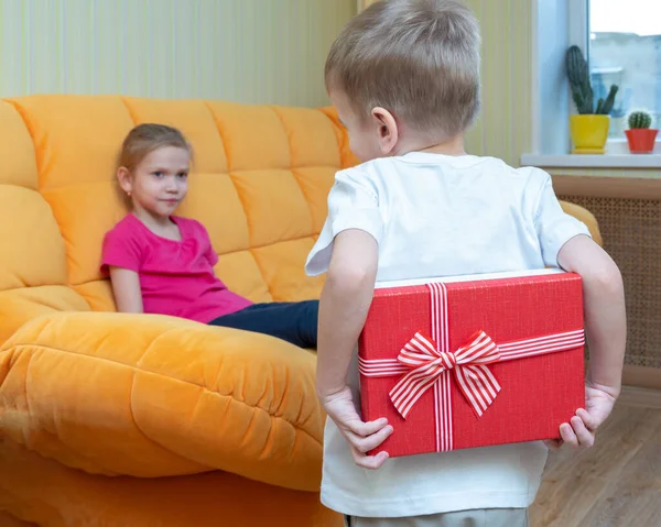 El momento de esperar un regalo. El chico sostiene una caja de regalo roja detrás de su espalda para darle a la chica sentada en el sofá. Feliz cumpleaños, feliz día de la hermana, feliz día de las niñas — Foto de Stock