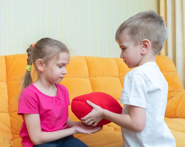 The moment of presenting the gift. A boy giving a red pillow in the shape of a heart to a girl sitting on the couch. Happy birthday, happy sister day, happy girls day — Stock Photo, Image