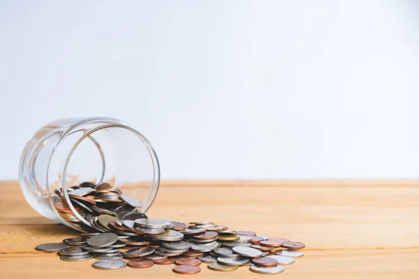 Coins in glass jar falling with money scattered on brown wooden table surface against white background, money-saving concept for future perfect, failure concept of money saving, finance, and banking.