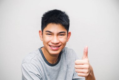 Genç Asyalı bir adamın portresi, başparmaklarıyla gülümseyen, mükemmelliğini ifade eden, ya da başarılı jestler yapan. Beyaz duvar arka planında insanların yüz ifadeleri ve duyguları var..