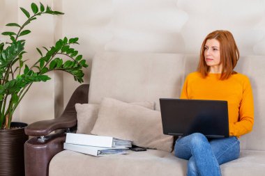 Güzel bir kadın dizüstü bilgisayarla kanepede oturuyor ve evden çalışıyor.