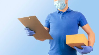 Mavi tişörtlü teslimatçı üniformalı steril yüz maskesi eldivenli, elinde karton kutular ve mavi arka planda izole tablet olan. Kurye servisinde çalışan adam karantina pandemik koronavirüs virüsü konsepti