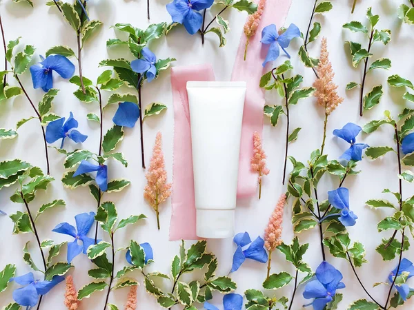 Mock-up of white squeeze bottle plastic cosmetic tube, fresh greens, blue and pink flowers, silk ribbons on white background. Bottle for branding and label. Natural organic spa cosmetics concept.