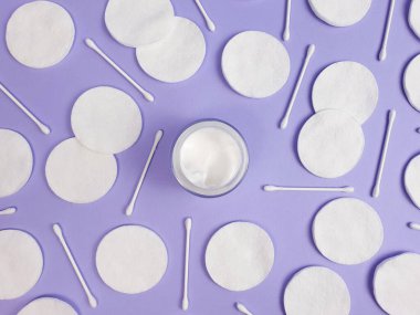 Açık cam kavanozda nemlendirici krem, pamuk pedler ve desenli açık mor arka planda pamuk çubuklar. Üst Manzara. Doğal biyoorganik eko cilt bakım ürünü.