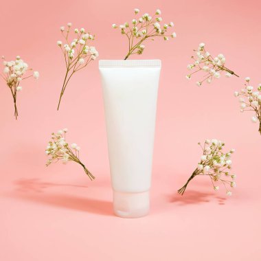 Beyaz plastik tüp ve açık pembe arka planda uçan çingene çiçeklerinin üst görüntüsü. Doğal organik spa kozmetik konsepti. Ön görünüm