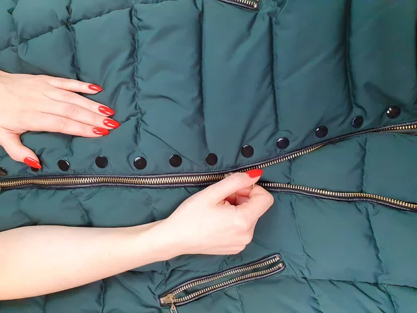 Female hands with red nails close zipper lock on down jacket. Autumn jacket fabric background, green puffer jacket texture. Top view