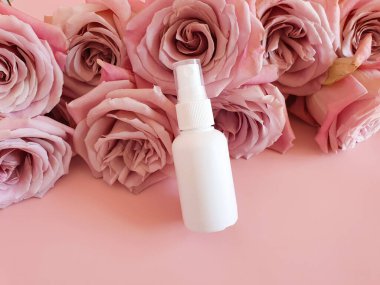 Markasız beyaz kozmetik sprey şişesi ve pembe arka planda bir sürü pembe gül. Modelleme. Cilt nemlendirici, sıvı antibakteriyel sprey konsepti. Mini seyahat versiyonu. Doğal vücut sisi.