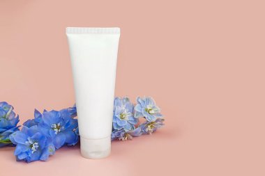 Damgasız beyaz sıkılmış krem tüpü ve pembe arka planda mavi Delphinium çiçeği. İlaç ya da kozmetik ürünleri için kozmetik tüp. Krem, jöle, cilt bakımı. Model, yakın çekim, ön görünüm.
