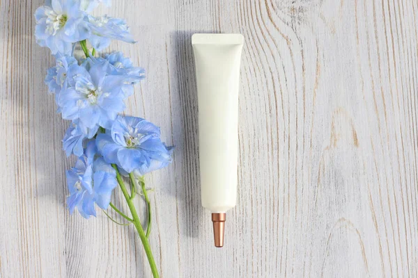 Face and eye cream squeeze cosmetic tube with long nozzle and bronze screw cap and blue Delphinium flower on white wood table. Natural organic cosmetics concept, flatlay, top view. Mockup, template.
