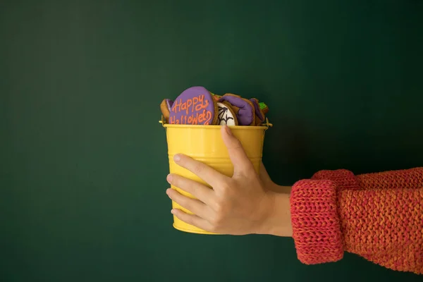 Child`s hands hold out a bucket filled with gingerbreads in the form of pumpkins. Concept of sweet gifts for Halloween party. Dark green background. Copy space