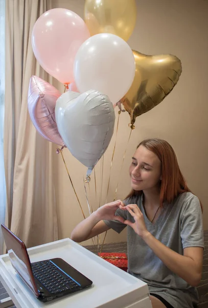 Kızıl saçlı genç bir kız arkadaşlarıyla görüntülü konuşma yapıyor ve onlara hediye ve balonlar için teşekkür ediyor. Koronavirüs salgını sırasında sosyal uzaklık ve kendini izole etme kavramı