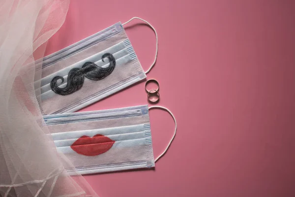 stock image Two wedding rings and protective face masks with a mustache and lips painted on them are on pink background. The concept of wedding ceremonies during a pandemic of coronavirus COVID-19