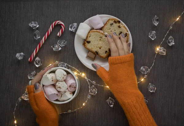 Girl\'s hands in orange knitted mittens hold a mug of hot drink with marshmallow and lollipop on a table with plate with pieces of cake and chocolate, decorated with shining garland. Christmas atmosphere