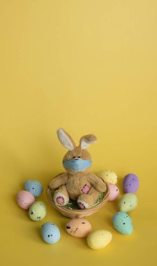 Yuvasında koruyucu yüz maskesi içinde tüylü oyuncak tavşan ve sarı arka planda çok renkli yumurtalar. Coronavirus Covid-19 salgını sırasında Paskalya tatili. Metnin için kopyalama alanı olan dikey pankart