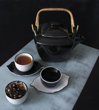 Güzel siyah ve beyaz çay seremonisi seti: çaydanlık, küçük fincanlar ve çay fincanları, siyah ahşap bir masanın üzerindeki mermer bir paspasın üzerinde kurumuş çay yapraklarıyla dolu bir fincanın yanında.