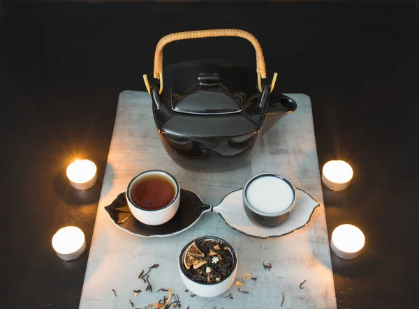 Beautiful black and white tea ceremony set: teapot and small cups with tea and milk and saucers next to a cup filled with dried tea leaves with pieces of citrus on the background of a black table with burning candles