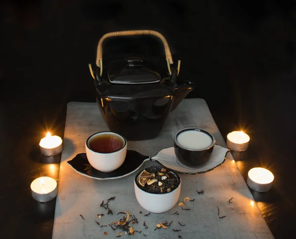 Beautiful black and white tea ceremony set: teapot and small cups with tea and milk and saucers next to a cup filled with dried tea leaves with pieces of citrus on the background of a black table with burning candles