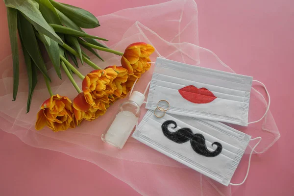 Two disposable protective masks with painted mustache and lips, two wedding rings and sanitizer next to bouquet of tulips on white veil on pink background. The wedding ceremony during COVID-19.