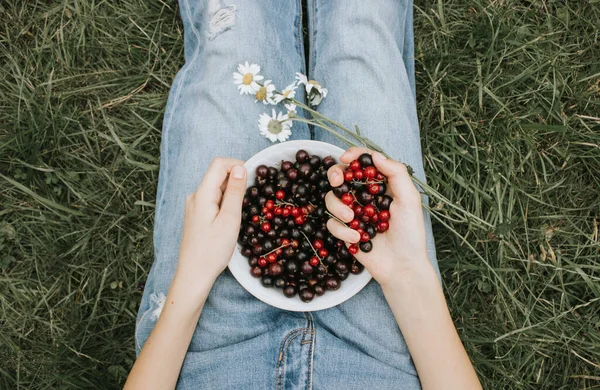 ジーンズの女の子が草の上に座り 庭からジューシーで香り高い黒と赤のスグリでプレートを持っています 幸せな子供時代 ビタミンが豊富な健康食品の概念 — ストック写真
