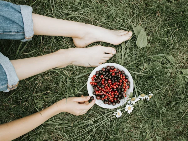 ジーンズの女の子が草の上に座り 庭からジューシーで香り高い黒と赤のスグリでプレートを持っています 幸せな子供時代 ビタミンが豊富な健康食品の概念 — ストック写真