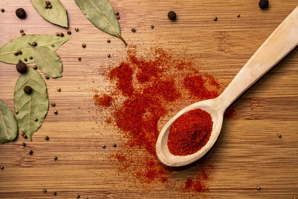 Ahşap bir arka planda kırmızı biber kaşığı. Yukarıdan bak. Arka planda sarımsak ve defne yaprağı var. Et ve baharatlı yemekler için baharatlar..
