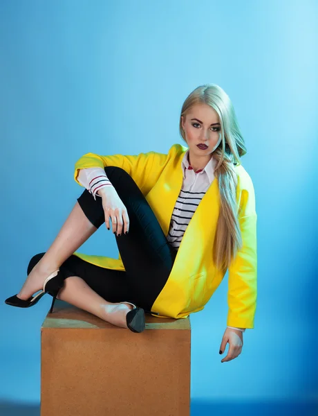 Portrait of blond model in fashion yellow coat sitting over blue — Stock Photo, Image