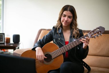 Kız oturma odasında akustik gitar çalıyor. Evde internetten ders alıyor.