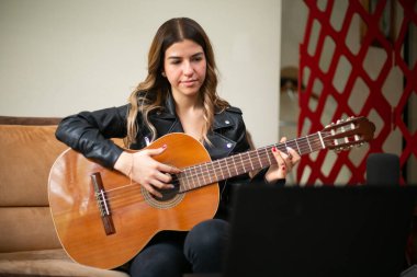 Kız oturma odasında akustik gitar çalıyor. Evde internetten ders alıyor.