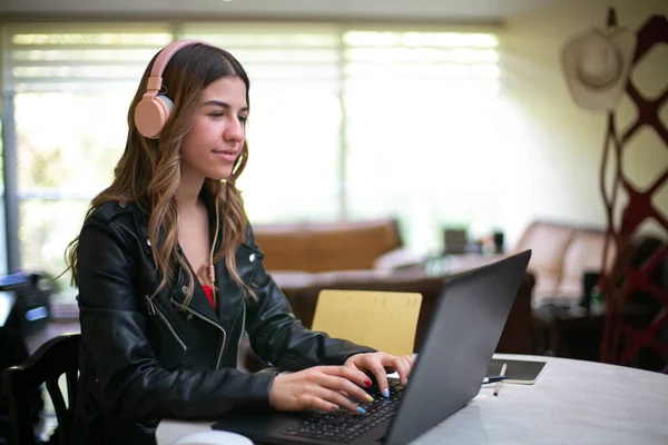 Young woman sitting in front of her laptop, she is doing her homework. She has a smiling expression and is looking at her laptop. She wears pink headphones