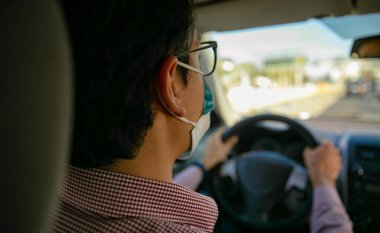 Yetişkin bir adam, kırmızı gömlekli, arabasını kullanıyor. Maskesi var.