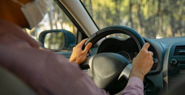 Yetişkin bir adam, kırmızı gömlekli, arabasını kullanıyor. Maskesi var.