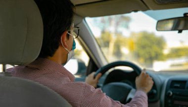 Yetişkin bir adam, kırmızı gömlekli, arabasını kullanıyor. Maskesi var.