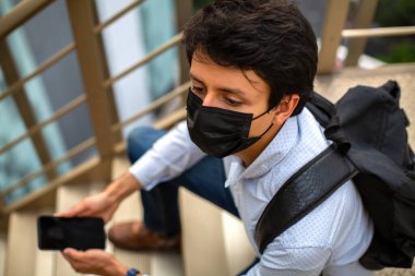 Maskeli genç adam, yemek merdivenlerinde oturuyor, telefonu elinde.
