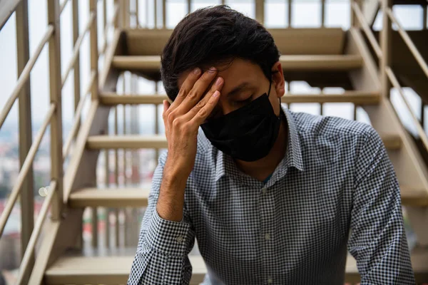 Young Men with mask, he is sitting on meal stairs, He has his hands in the mask and he is worried.