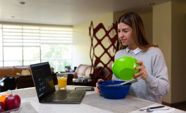 Genç kadın laptopuyla yemek yapmayı öğreniyor. O gülümsüyor. Mutfakta..