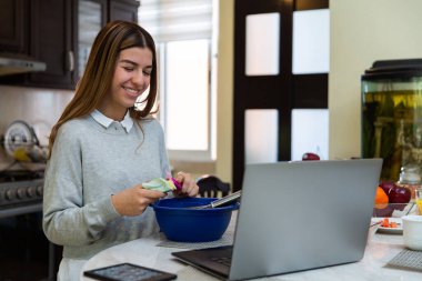 Genç kadın laptopuyla yemek yapmayı öğreniyor. O gülümsüyor. Mutfakta..