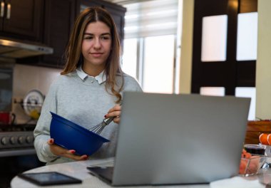 Genç kadın laptopuyla yemek yapmayı öğreniyor. O gülümsüyor. Mutfakta..