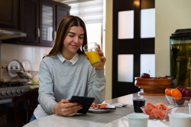 Gülümseyen genç bir kadın evdeki mutfak masasında sağlıklı bir kahvaltı yaparken dijital tabletten bir şeyler izliyor.