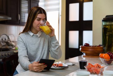 Gülümseyen genç bir kadın evdeki mutfak masasında sağlıklı bir kahvaltı yaparken dijital tabletten bir şeyler izliyor.