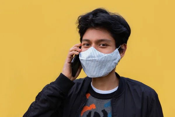 Maskeli genç latin adam telefonunu kullanıyor. Sarı arkaplan